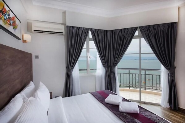 A hotel room with a large bed, a view of the sea through floor-to-ceiling windows, and a balcony with a railing.