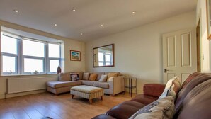 Ein Wohnzimmer mit Ecksofa, Pouf, Beistelltisch und großem Fenster.