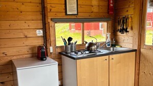A compact kitchen with wooden cabinets, a white refrigerator, a sink, and a window with a view of a red building outside. - Hólaskjól Highlandcenter (Kirkjubaejarklaustur)