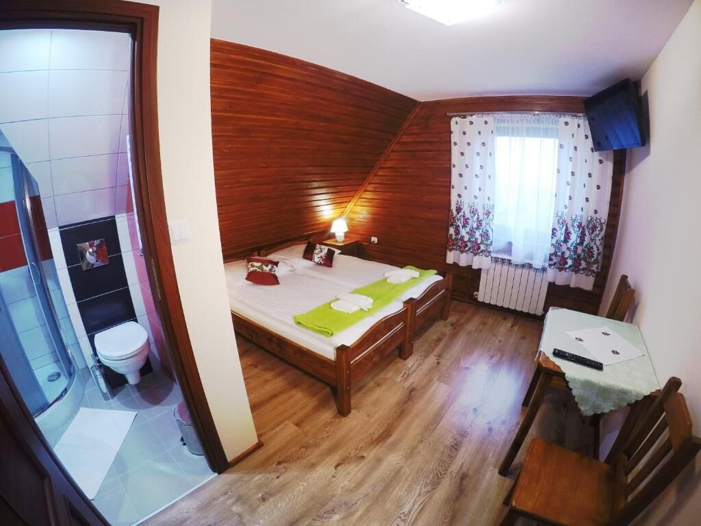 A bedroom with a wooden ceiling, a bed with green towels, a bedside table, a TV, and a window with floral curtains.