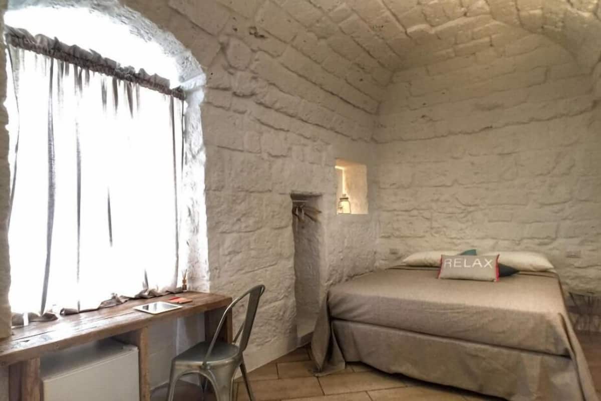 Una habitación rústica con pared de piedra, una cama con almohada que dice 'RELAX', un escritorio con silla y una ventana pequeña.