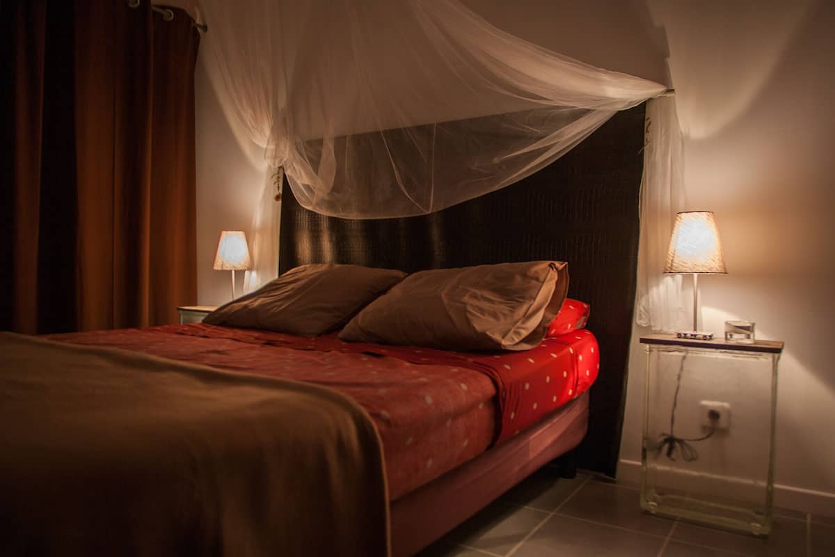Un dormitorio con cama, mesita de noche, lámpara y cortina.
