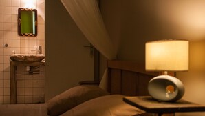 A dimly lit room with a bed, a bedside table with a lamp, a small wooden nightstand, and a bathroom with a sink and mirror. - Les deux chemins (Mons)