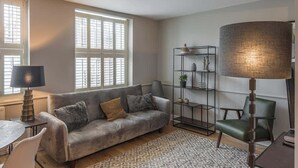 Ett vardagsrum med en soffa, en lampa, en fåtölj, en bokhylla och ett fönster med persienner.