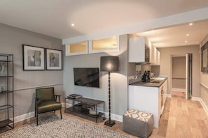 A modern living room with a sofa, a coffee table, a floor lamp, and a TV. There is a kitchen area with cabinets and a countertop.