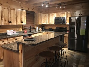 A kitchen with wooden cabinets, granite countertops, a stainless steel refrigerator, a microwave, and a stove. There are two stools at the island. - Beautiful 3 bed / 3 bath Luxury Cabin, Hot Tub, Games, Easy Check in! Book Now! (Sevierville)
