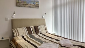 En pænt redt seng med stribet sengetøj og to hvide håndklæder, et billede af strandhuse på væggen, og et vindue med lodrette persienner.