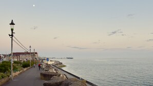 En kystpromenade med en bro i baggrunden, klar himmel og et par personer der går.