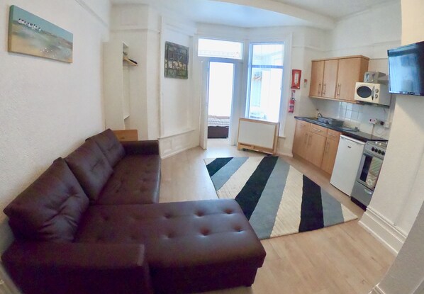 A living room with a dark brown sofa, a kitchen with wooden cabinets, a microwave, and a stove. - Cozy apartment for 4 in fashionable North Shore (Blackpool)