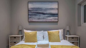 Una cama bien hecha con una toalla doblada, dos mesitas de noche con lámparas, y un cuadro de un paisaje en la pared.
