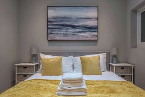 A neatly made bed with a folded towel, two bedside tables with lamps, and a painting of a landscape on the wall. - Nelson West- Modern one bed, great for long stays (Ramsgate)