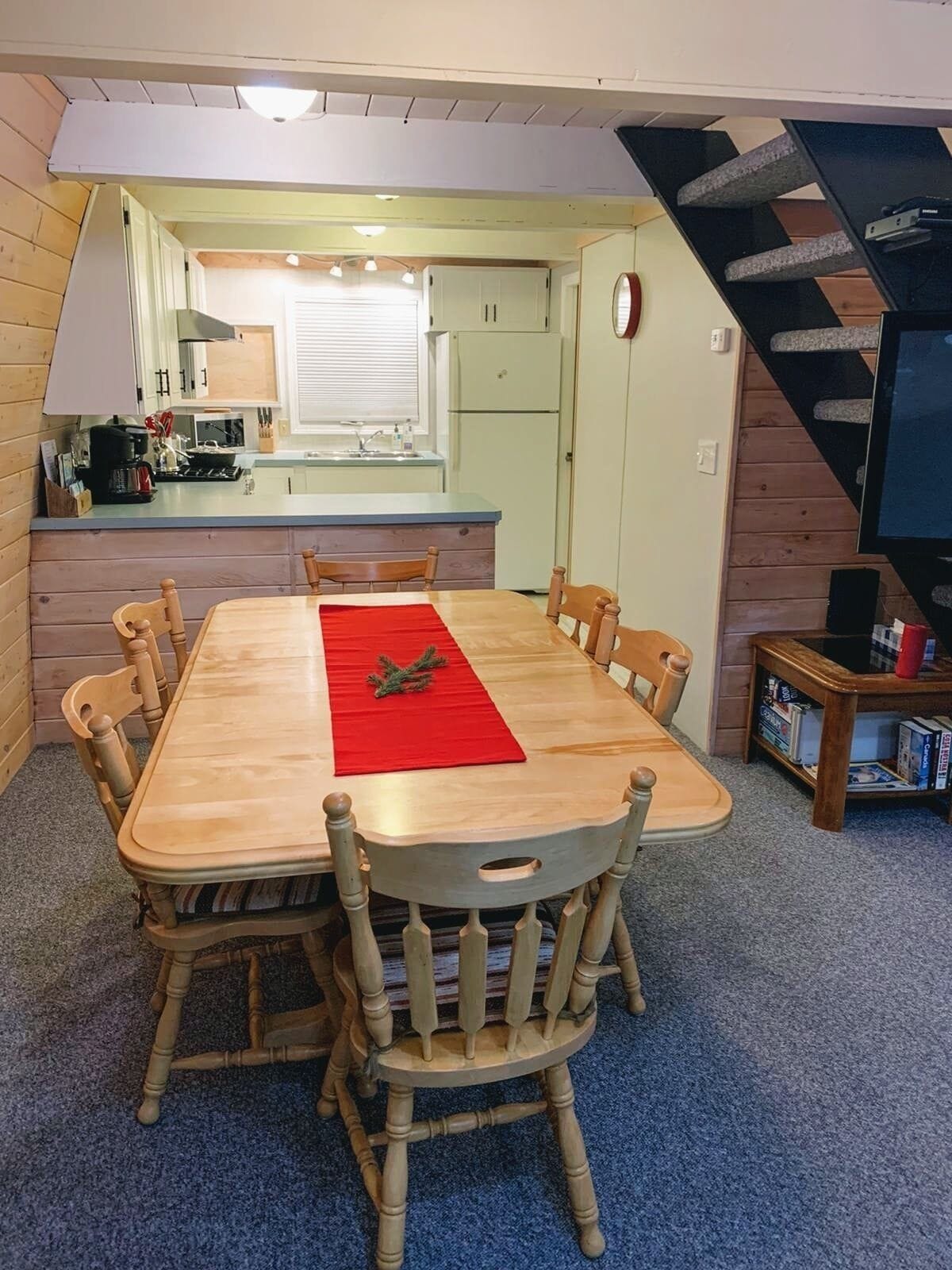 A dining area with a wooden table and chairs, a red runner, and a kitchen in the background.