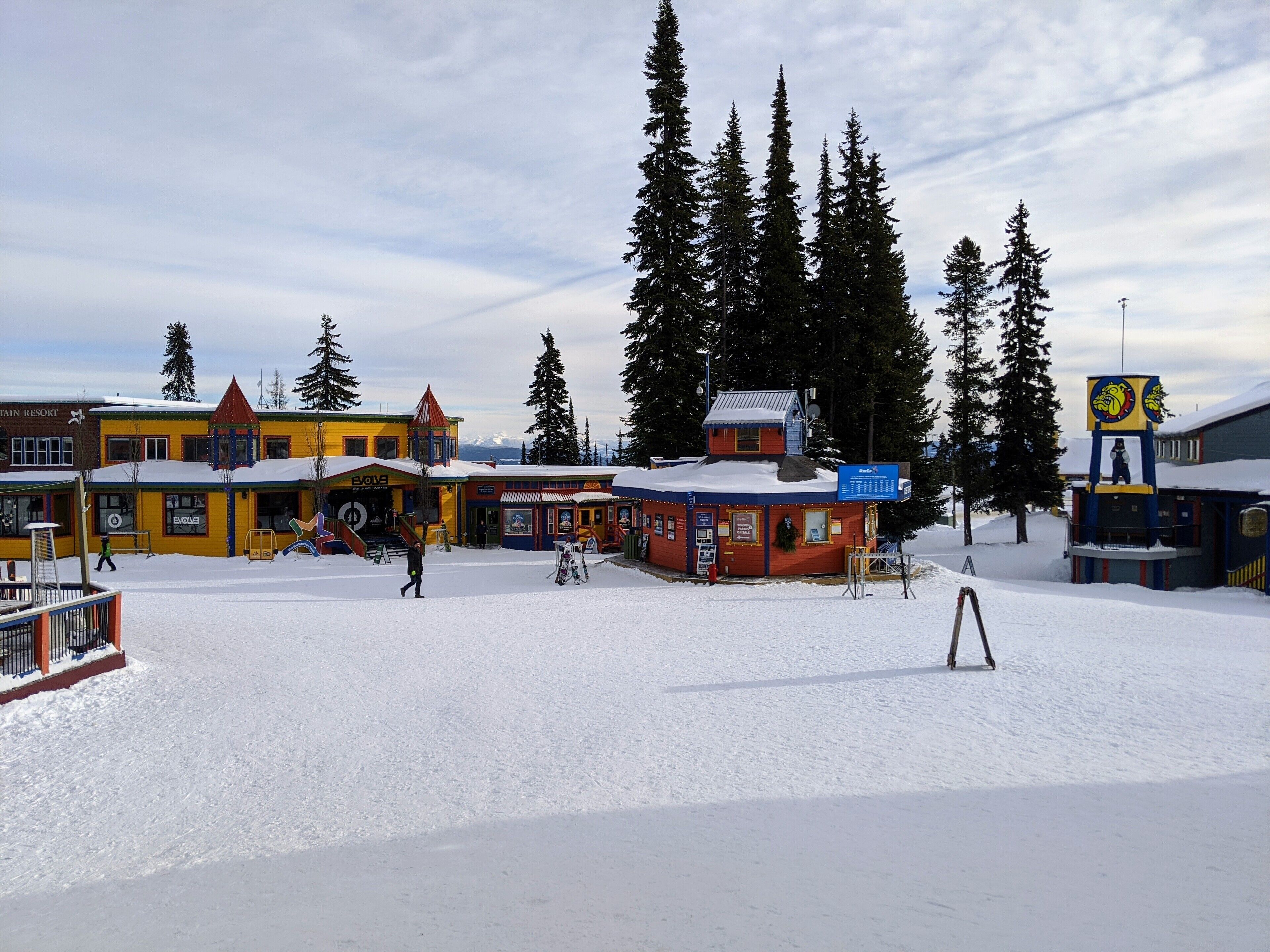Ett snötäckt utomhusområde med byggnader, inklusive en gul byggnad med rött tak och en blå byggnad med en skylt.
