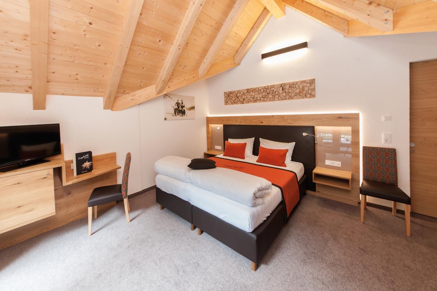 A modern bedroom with a wooden ceiling, a bed with orange and white bedding, a TV, and a desk with a chair.