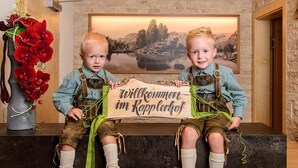 Dos niños con atuendo tradicional sostienen un letrero de bienvenida en una habitación con pared de piedra y un cuadro de un paisaje.