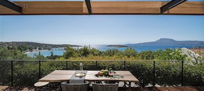 A wooden table with a glass on it, surrounded by wicker chairs, overlooking a coastal view with a clear sky and distant mountains.