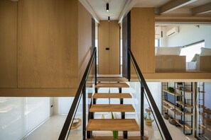 A modern interior with a wooden staircase, glass railings, and a view of a bedroom with a bed and a TV.