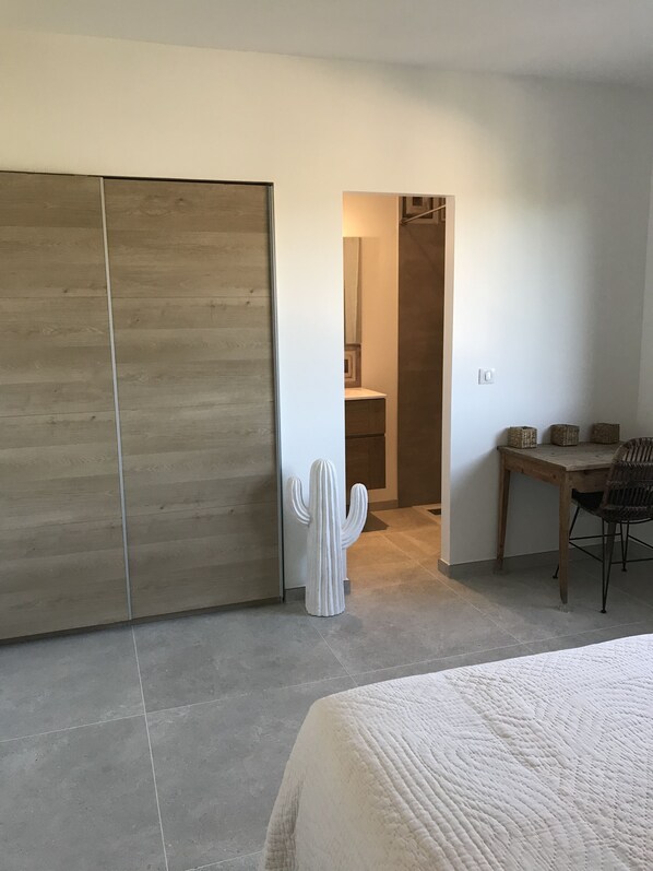 A bedroom with a bed, a wooden wardrobe, a cactus sculpture, a dining table with chairs, and a small kitchen area.