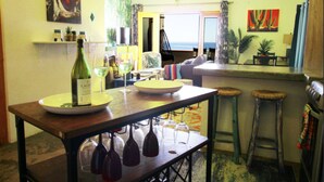 A dining area with a wooden table, wine glasses, and a bottle of wine. There are two stools and a chair. The room has a view of the ocean through a window.