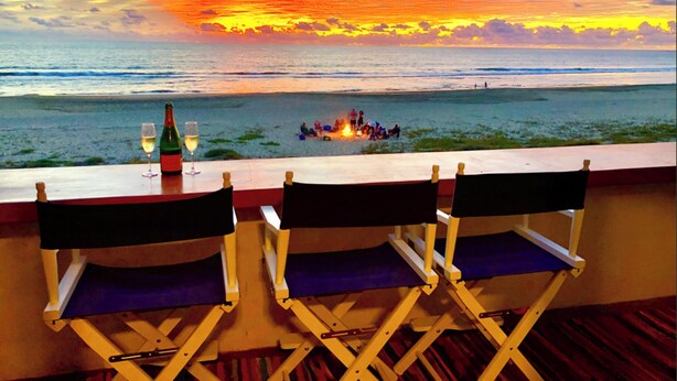 A balcony with a view of the sunset over the ocean, a bottle of wine, and two folding chairs.