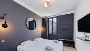 A modern hotel room with a large bed, a round mirror, a TV mounted on the wall, and a view of the city through the window. - Heart of Cannes Amazing design apt Croisette Top Location Olam Properties (Cannes)