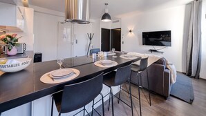 A modern kitchen with a central island, a dining area with chairs, a sofa, and a TV mounted on the wall. - Heart of Cannes Amazing design apt Croisette Top Location Olam Properties (Cannes)