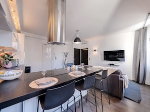 A modern kitchen with a central island, a dining area with chairs, a sofa, and a TV mounted on the wall. - Heart of Cannes Amazing design apt Croisette Top Location Olam Properties (Cannes)