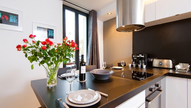 A modern kitchen with a dining area, featuring a central island with a vase of red flowers, a wine bottle, and dining utensils. - Heart of Cannes Amazing design apt Croisette Top Location Olam Properties (Cannes)