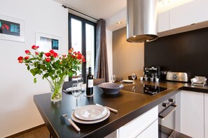 A modern kitchen with a dining area, featuring a central island with a vase of red flowers, a wine bottle, and dining utensils. - Heart of Cannes Amazing design apt Croisette Top Location Olam Properties (Cannes)