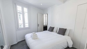 Un lit bien fait, avec du linge de lit blanc et une pile de serviettes, une fenêtre avec des stores et une porte donnant sur une autre pièce.