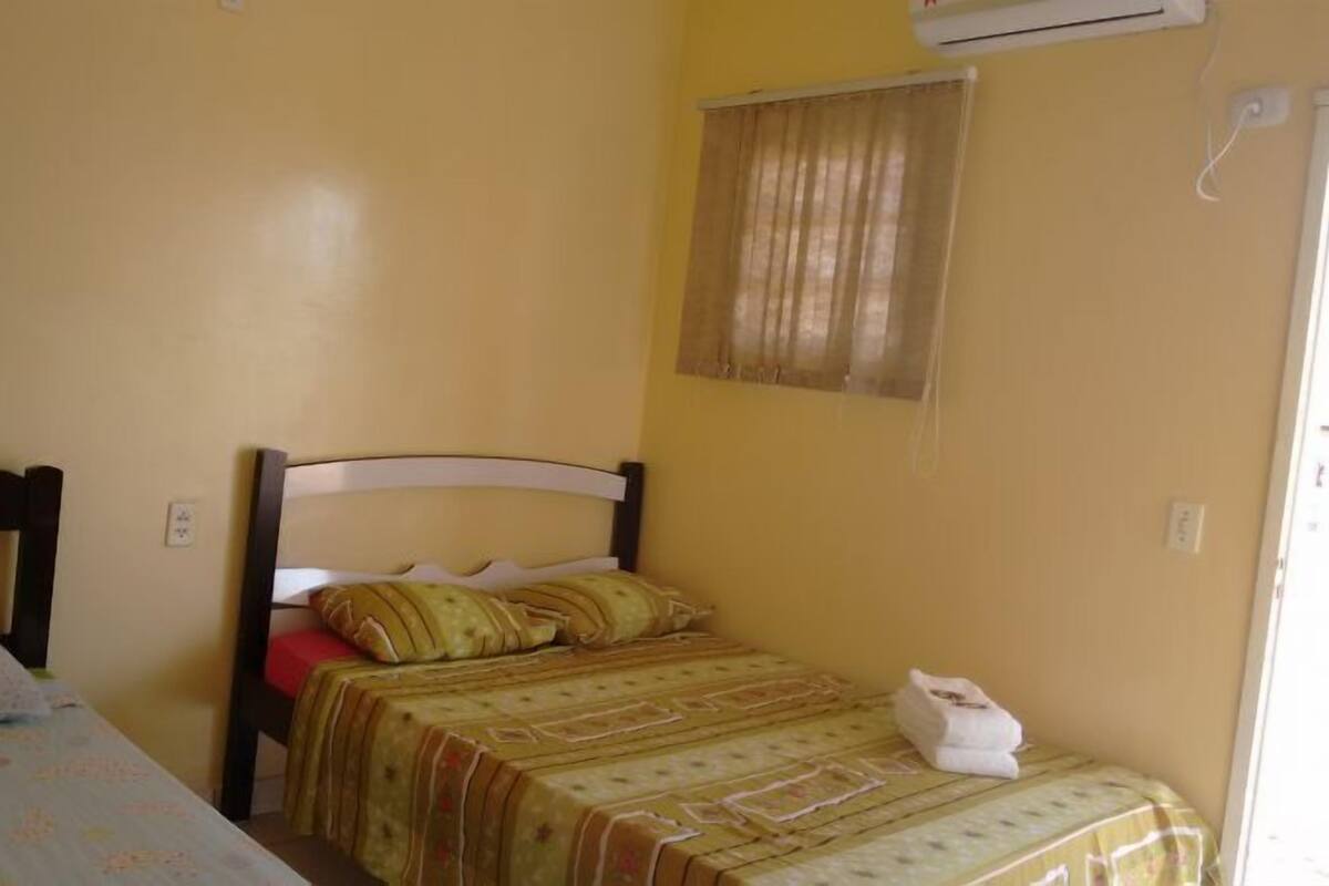 Une petite chambre avec un lit simple, une tête de lit en bois, une fenêtre avec des rideaux et un appareil de climatisation fixé au mur.