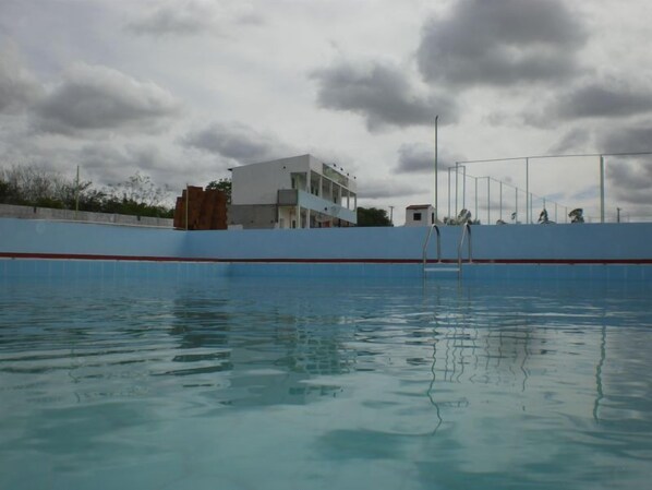 Una piscina con borde de azulejos azules y blancos, un edificio blanco con una franja roja al fondo y un cielo nublado.