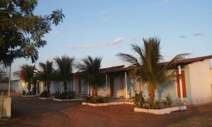 A row of single-story buildings with a dirt ground, surrounded by palm trees. - Pousada Nathymar (Piranhas)