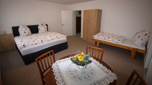 A bedroom with a double bed, a single bed, a wooden wardrobe, a table with a lace tablecloth, and a chair.