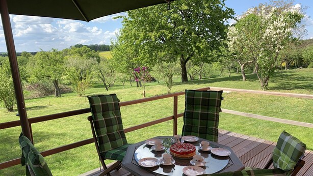 A deck with a table set for tea, surrounded by chairs with plaid cushions, overlooking a grassy landscape with trees.