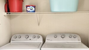 Ein Waschbereich mit zwei Whirlpool-Waschmaschinen und einem darüberliegenden Regal, das einen roten Topf, eine blaue Schüssel und einen Reiniger enthält.