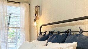 Ein ordentlich bezogenes Bett mit weißen und dunkelblauen Kissen, einem Metall-Kopfschild und einem Fenster mit transparenten Vorhängen.