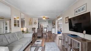 A spacious living room with a gray sofa, a wooden coffee table, and a dining area with wicker chairs. A kitchen with white cabinets and a large TV is visible in the background.