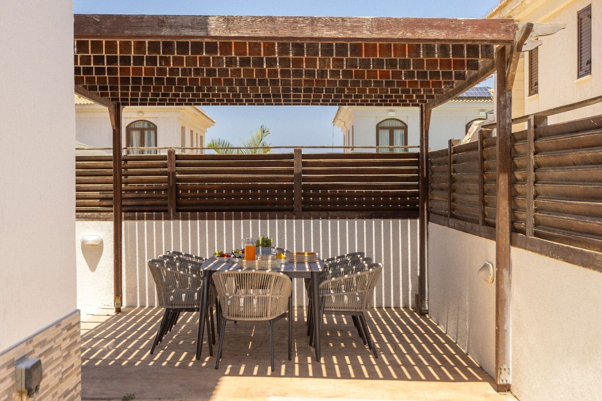 Une terrasse extérieure dotée d’une pergola en briques, d’une table avec des chaises et offrant une vue sur les bâtiments en arrière-plan.