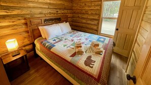 A wooden cabin bedroom with a bed, a nightstand, a lamp, and a door. - Secluded Creekside Cabin (Coaldale)