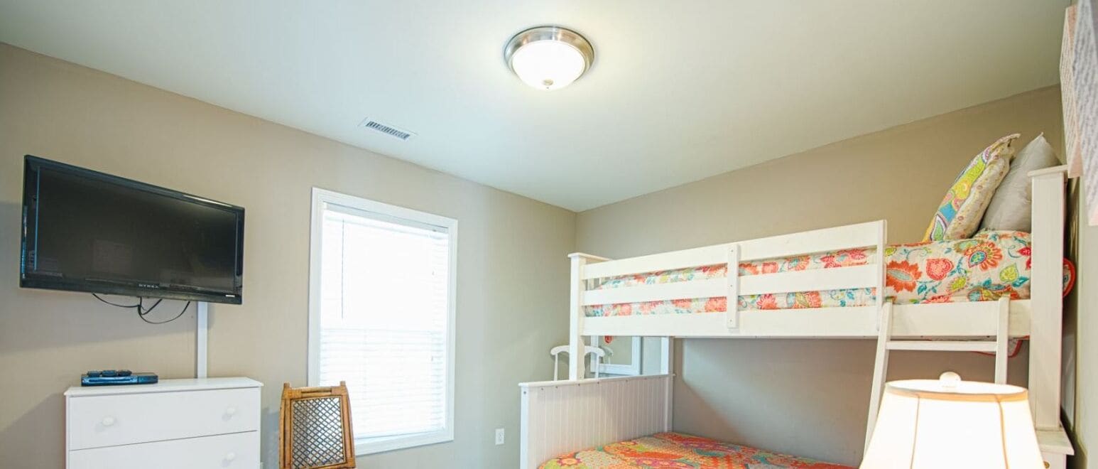 A bedroom with a bunk bed, a television, a desk with drawers, a chair, and a window with blinds.