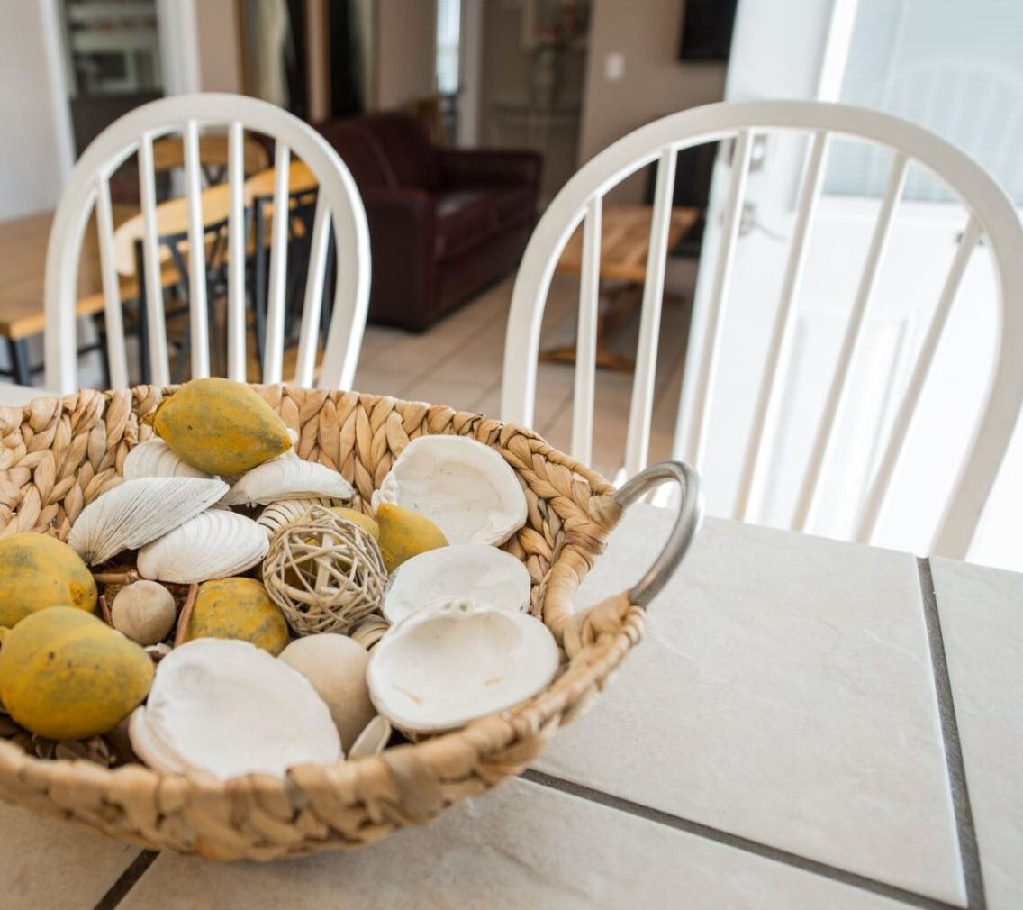 A wicker basket filled with seashells and a decorative ball on a white table in a room with white chairs.