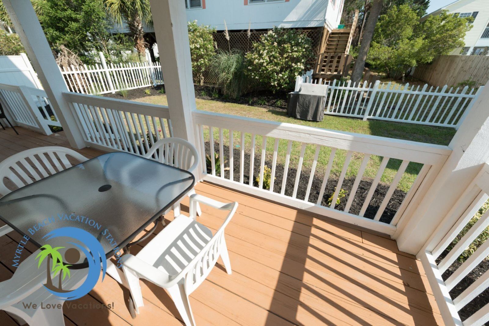 A covered outdoor area with a table and chairs, surrounded by white picket fences and greenery.