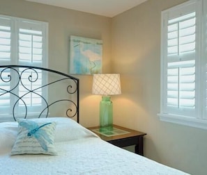 A bedroom with a bed, a nightstand, a lamp, and a window with shutters. - Windy Hill Villa 4 - Beach Fun (North Myrtle Beach)