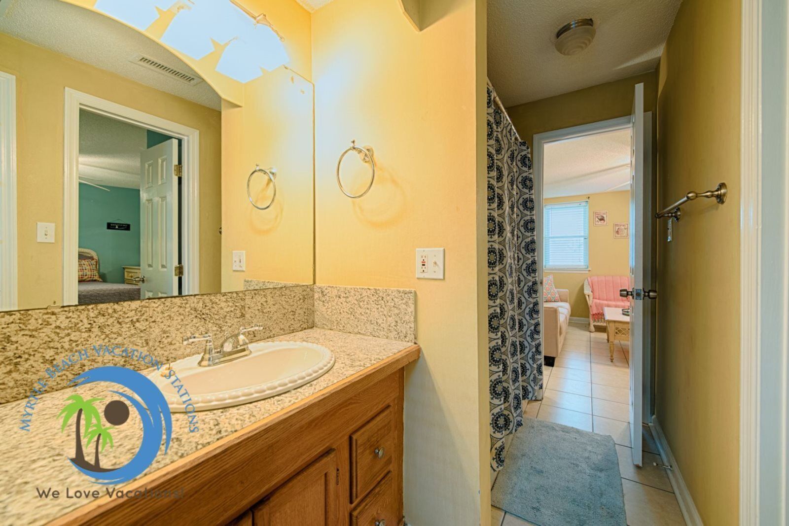 A bathroom with a granite countertop, a sink, and a mirror. There is a door leading to a bedroom with a bed and a window.
