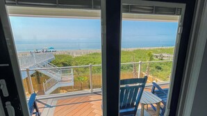 A view from a room with a sliding glass door to a deck with outdoor furniture and a beach in the background. - "Be Still" - Oceanfront Home w/ Private Pool, 4 Bedrooms / 3.5 Bathrooms (Holden Beach)
