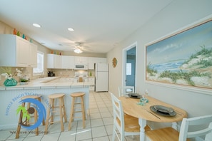 A kitchen with white cabinets, a wooden island with stools, a painting of a beach scene, and a ceiling fan.