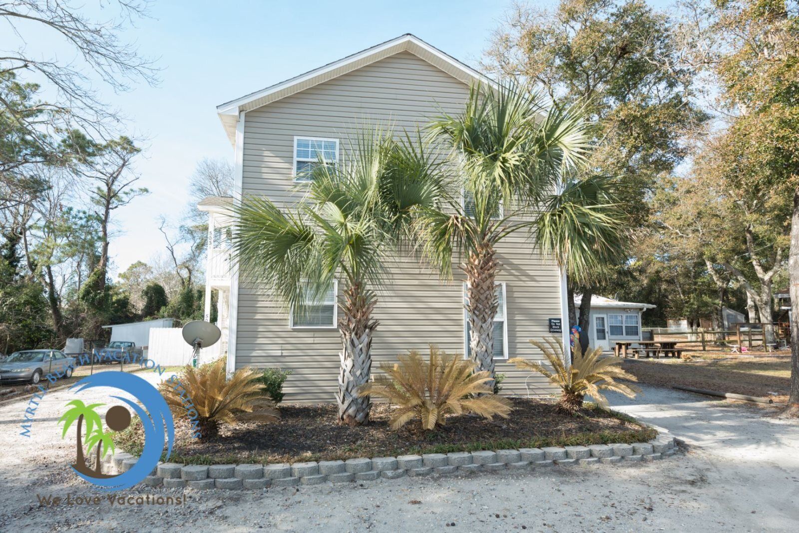 A single-story house with a beige exterior, surrounded by palm trees and a gravel driveway.