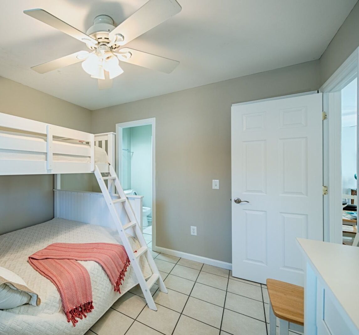 A bedroom with bunk beds, a ladder, a ceiling fan, a door, and a small desk with a chair.