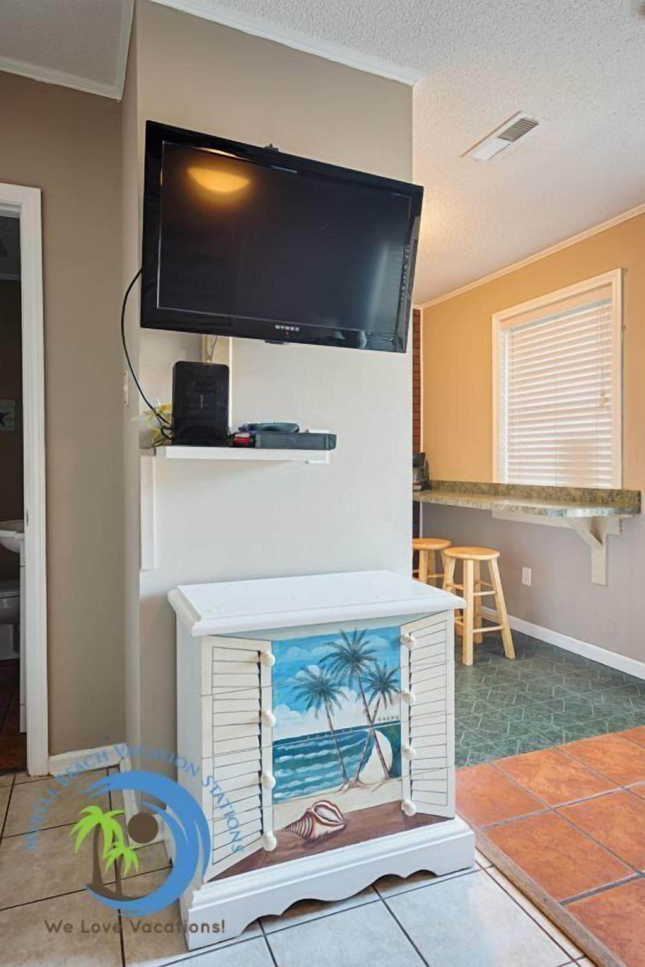 Un salon avec un téléviseur fixé au mur, un meuble de rangement blanc au design inspiré du bord de mer, et un coin cuisine avec une fenêtre et des tabourets de bar.